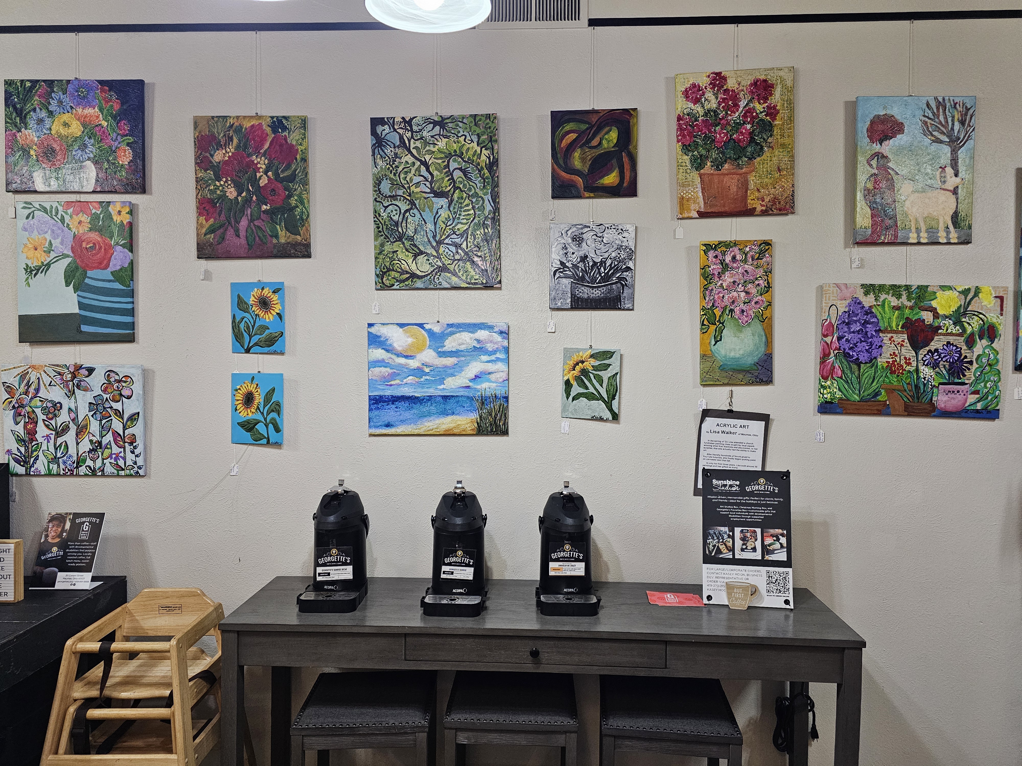 Wall display of colorful, handcrafted paintings by local artists above a coffee station, featuring floral, landscape, and abstract pieces arranged in a community gallery setting.